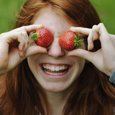 Hymyilevä tyttö pitää sormillaan kahta mansikkaa silmiensä edessä.