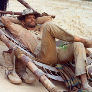 Hymyilevä lännenasuinen mies (näyttelijä Terence Hill) makailee hevosen valjaisiin kiinnitetyllä kenttävuoteella tai paarilla jalka toisen polven päällä, kädet ristissä pään takana ja hattu silmillä. Kuva elokuvasta Trinity ratsastaa jälleen.