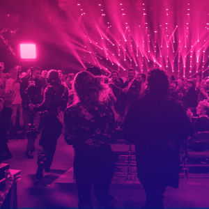 Människor som står upp och klappar i händerna i en konsertlokal med ett lila-rosa sken.