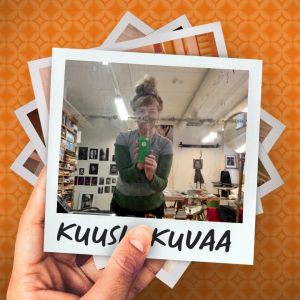 Taitelija Ulla Jokisalo työhuoneellaan kuluneen peilin kautta otetussa selfiessä. Etualalla työpöytä, takana seinällä valokuvia.