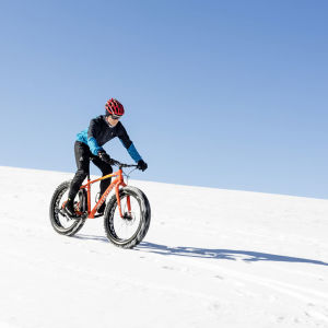 Pyöräilijä ajaa alas lumista rinnettä. Taustalla sininen aurinkoinen taivas.