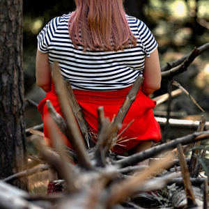 Nainen istuu kaatuneen puun päällä selkä kameraan päin. Puusta törröttää teräviä oksia.