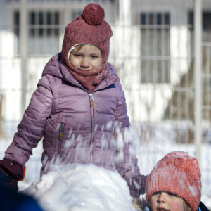 Kaksi pipopäistä lasta ulkona, toinen heittää lunta lumikasan päälle.
