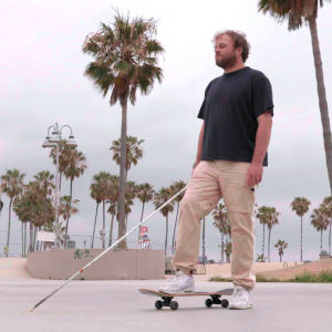 Brian Bushway seisoo betonisella aukiolla toinen jalka rullalaudan päällä kädessään valkoinen keppi. Taustalla on Venice Beach ja palmupuita.