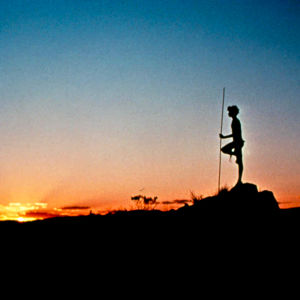 Nuori mies lannevaatteessa seisoo profiilissa aavikolla vasten auringonlaskua. Kuva elokuvasta Walkabout.