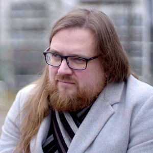En man med långt hår, skägg och glasögon betraktar lugnt något utanför bild.