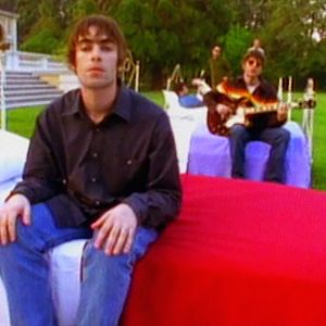 Mies istuu sängyllä, jossa punainen peitto. Taustalla toinen mies istuu toisella sängyllä, jossa violetti peitto, ja soittaa kitaraa.