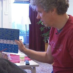 En person håller upp en skylt med bokstäver och ord på, medan en äldre person i rullstol pekar på symbolerna för att kommunicera. 