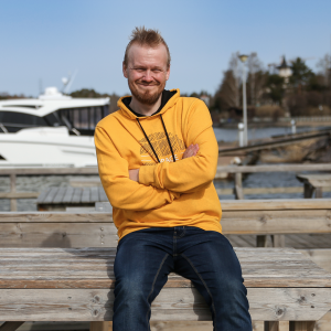 Jyrki Hakanen istumassa rannalla, vene taustalla.