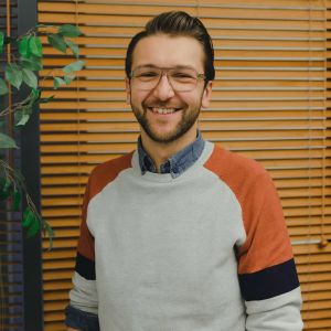 Sebastian småler mot kameran. Han står mot en skärmgardin och på vänstra sidan syns en grönväxt.