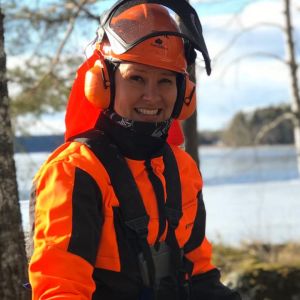 Arja Koriseva hymyilee kameralle metsämaisemassa metsurivarusteissa ja raivaussaha käsissään.