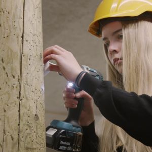 De flesta håller fokus på att ta sin yrkesexamen. Fördröjningar kan det bli men det verkar inte bli fler som avbryter helt. Ung kvinna i förd gul skyddshjälm håller i en elektrisk borr och borrar i en kraftig trästolpe med fokuserad blick.