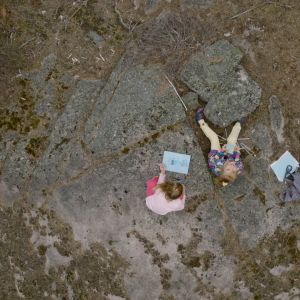 Kaksi tyttöä istuu kalliolla, kuva on kuvattu ylhäältä päin.