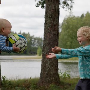 Pieni lapsi pitää palloa sylissään ja isompi lapsi ojentaa käsiään palloa kohti.