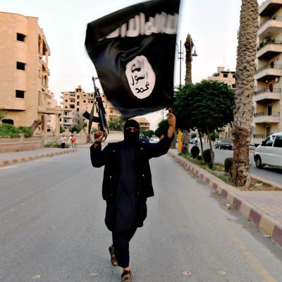 Mustiin pukeutunut aseistaunut henkilö pitelee ISIS-lippua ilmassa.