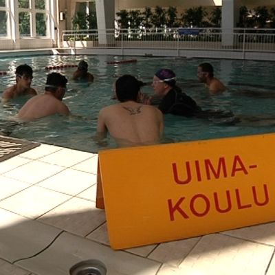 Maahanmuuttajia uimakoulussa. Uimakoulukyltti uima-altaan reunalla.