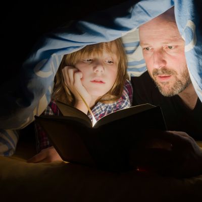 Isä ja poika lukevat kirjaa peiton alla taskulampun valossa