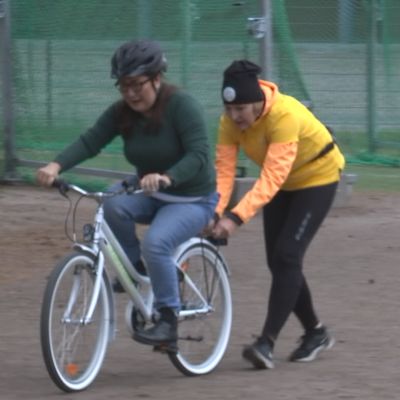 Nainen ajaa pyörällä, jota toinen nainen tukee.