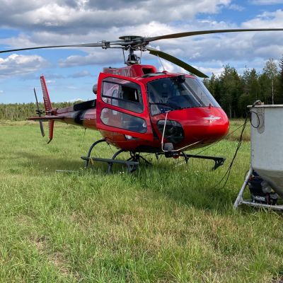 Punainen helikopteri pellolla, edessä valkoinen lannoitussäiliö.