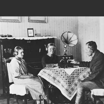 Radionkuuntelua 1920-luvulla.Kaksi naista ja mies pöydän ääressä kuuntelemassa radiota (kuulokkeet).