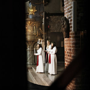 Bild tagen på långt håll. Luciatrio står framför ett altare, i bilden syns också män och tv-kamera.