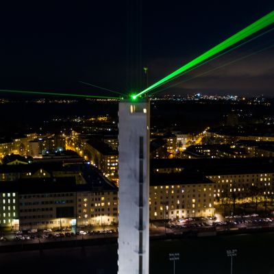 Stadionin torni pimeää taivasta vasten, vihreitä lasersäteitä ja kaupungin valoja. Valoshow sykkii läpi yön, ja morsettavat taivaalle merkkisarjaa: ”2020–2021”.