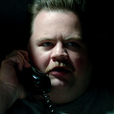 Närbild på Richard Jewell (Paul Walter Hauser) då han sitter i ett mörkt rum och håller i en telefonlur.