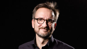 Vesa-Matti Lahti tycker att flygskatten är ett steg på vägen. Vesa-Matti Lahti, glasögonprydd med skägg och svart skjorta stående framför en svart bakgrund.