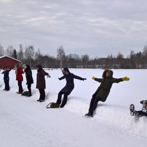 Noin kymmenen henkilöä seisoo lumisessa maisemassa tien reunassa, ja he oat kaatumassa taaksepäin selilleen pehmeään lumihankeen.