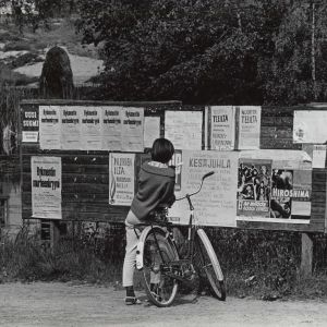 Maaseutu. Tyttö ja polkupyörä ilmoitustaulun edessä v. 1964. Elokuvamainoksia ja ilmoituksia. Kesä.