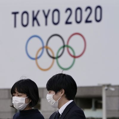 två personer står med ansiktsmasker framför en OS-skylt.
