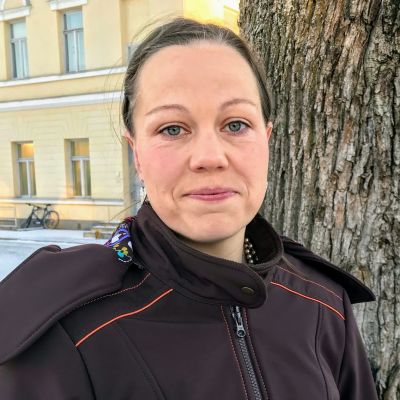 Sanna Timonen katsoo kameraa. Taustalla näkyy puunrunkoa ja keltainen rakennus.