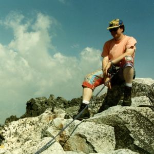 Vuorikiipeilyä harrastava mies istuu kalliolla. Hän on kiinni turvavaljaassa. Päässä on lippalakki.