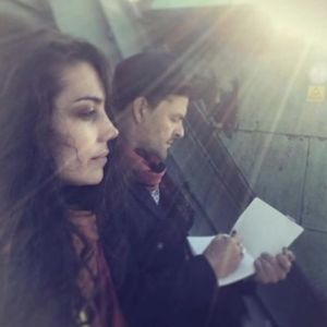 Nainen ja mies istuvat ilta-auringossa katolla. Sivukuvassa etualalla nainen, jolla on pitkät, kiharat hiukset. Hänen takanaan hattupäinen mies piirtää lehtiöön.