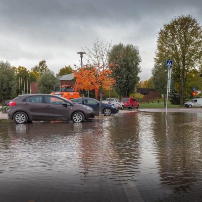 Bilar på en översvämmad parkeringsplats. Höstlandskap, en stor röd träbyggnad till höger. 