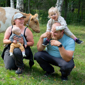 Iloinen pariskunta poseeraa kyykyssä, mukana myös vauva ja kaksivuotias, taustalla kulkee ohi valko-ruskea kyyttölehmä.