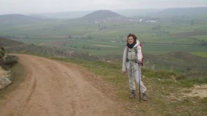 Santiago de Compostelan pyhiinvaellusreitillä tien reunassa avarassa maisemassa taustalla matalaa vuoristoa, Manuela Bosco seisoo vaellussauvaan nojaten ja katsoo kameraan.