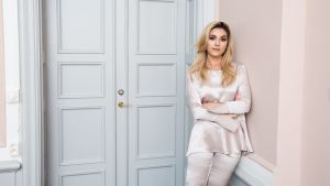 Det var psykologen som fick Michaela att inse att hon var utmattad. Michaela Forni iklädd en vit blus och vita byxor av siden. Hon står framför en stängd dörr, och lutar sig mot väggen bredvid.