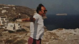 Leveästi hymyilevä italialaismies aurinkolaseissaan ja shortsiasussaan seisoo päivänpaisteessa rantakallioilla. Hän on levittänyt kätensä kuin lentoasentoon, taustalla näkyy yksinkertaisia kivitaloja ja merta sekä suuri laiva horisontissa.