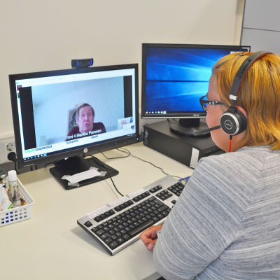 En kvinna sitter framför en dator. Hon har hörlurar på sig. På skärmen framför henne syns en äldre man som hon pratar med.
