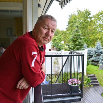 En man i röd tröja lutar sig över ett balkongräcke och tittar in i kameran.