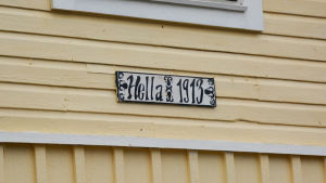 De flesta av de gamla trähusen är byggda i slutet av 1800-talet eller i början av 1900-talet. En skylt på en gul trähusvägg. På skylten står det Hella.
