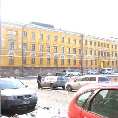 En gul stor byggnad på andra sidan ett torg. Det ligger snö på marken. Bilar står parkerade på gatan framför byggnaden.