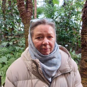 En kvinnlig författare som med en ljusblå duk på huvudet och en beige vinterkappa står sorgmodigt leende i Helsingfors Botaniska trädgård