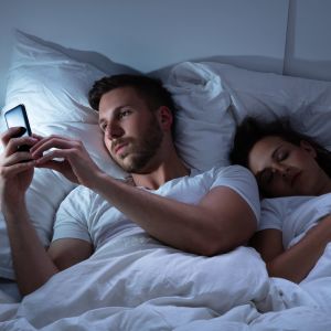 Man ligger i en säng och ser på en telefon, en kvinna sover bredvid honom