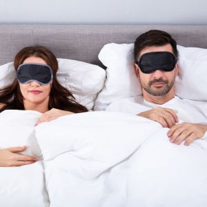Kvinna och man ligger i en säng, båda har svarta sov-masker över ögonen