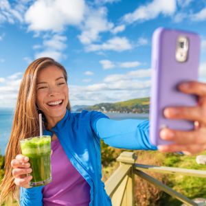 Leende ung kvinna tar en selfie framför en vacker vy och med en grön smoothie i handen