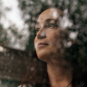 Katja Lahti seisoo sisällä kotonaan ja katsoo ulos ikkunasta, jossa on sateen jäljiltä vesipisaroita.