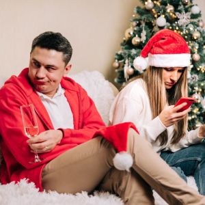 Man och kvinna i soffa framför julgran, hon tittar på sin telefon och han vänder sig irriterat bort