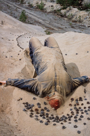 Haalaripukuinen henkilö makaa hiekassa käpyjen keskellä punainen huivi kasvojen suojana Turppi-ryhmän maataideteoksessa Muurahaiskuningatar Lehtimäessä 1982.